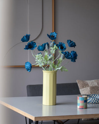 Een Kunstbloem Poppy van 73 cm in donkerblauw staat in een gele vaas op een tafel, vergezeld van een klein, kleurrijk gestreept kopje, in een moderne, zacht verlichte kamer met een minimalistische inrichting en neutrale tinten.