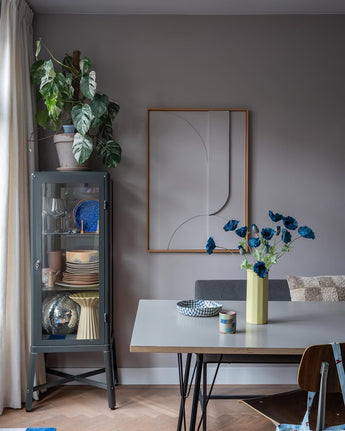 De moderne eethoek is voorzien van een grijze glazen vitrinekast waarin serviesgoed en planten zijn uitgestald, minimalistische wanddecoraties en een tafel waarop de Kunstbloem Poppy 73 cm donkerblauw in een gele vaas staat. De ruimte wordt gecompleteerd door een eclectische mix van stoelen, allemaal tegen een achtergrond van grijze muren en een houten vloer.