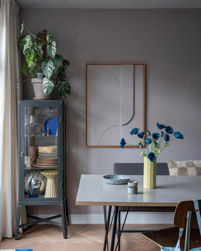 De moderne eethoek is voorzien van een grijze glazen vitrinekast waarin serviesgoed en planten zijn uitgestald, minimalistische wanddecoraties en een tafel waarop de Kunstbloem Poppy 73 cm donkerblauw in een gele vaas staat. De ruimte wordt gecompleteerd door een eclectische mix van stoelen, allemaal tegen een achtergrond van grijze muren en een houten vloer.