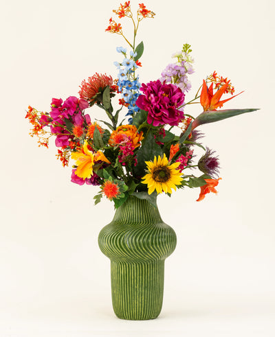 Het Zijden kunstbloemen boeket Colourful Spirit heeft levendige kunstbloemen, waaronder zonnebloemen en pioenrozen, gerangschikt in een hoge groene geribbelde keramieken vaas tegen een lichte achtergrond.