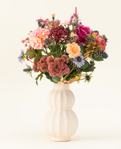 In een witte, gebogen vaas van keramiek staat het Zijden kunstbloemen boeket Pretty Pastel - een kleurrijke mix van duurzame bloemen zoals roze rozen, dahlia's, paarse en oranje bloemen en groen - prachtig gerangschikt tegen een lichte achtergrond.
