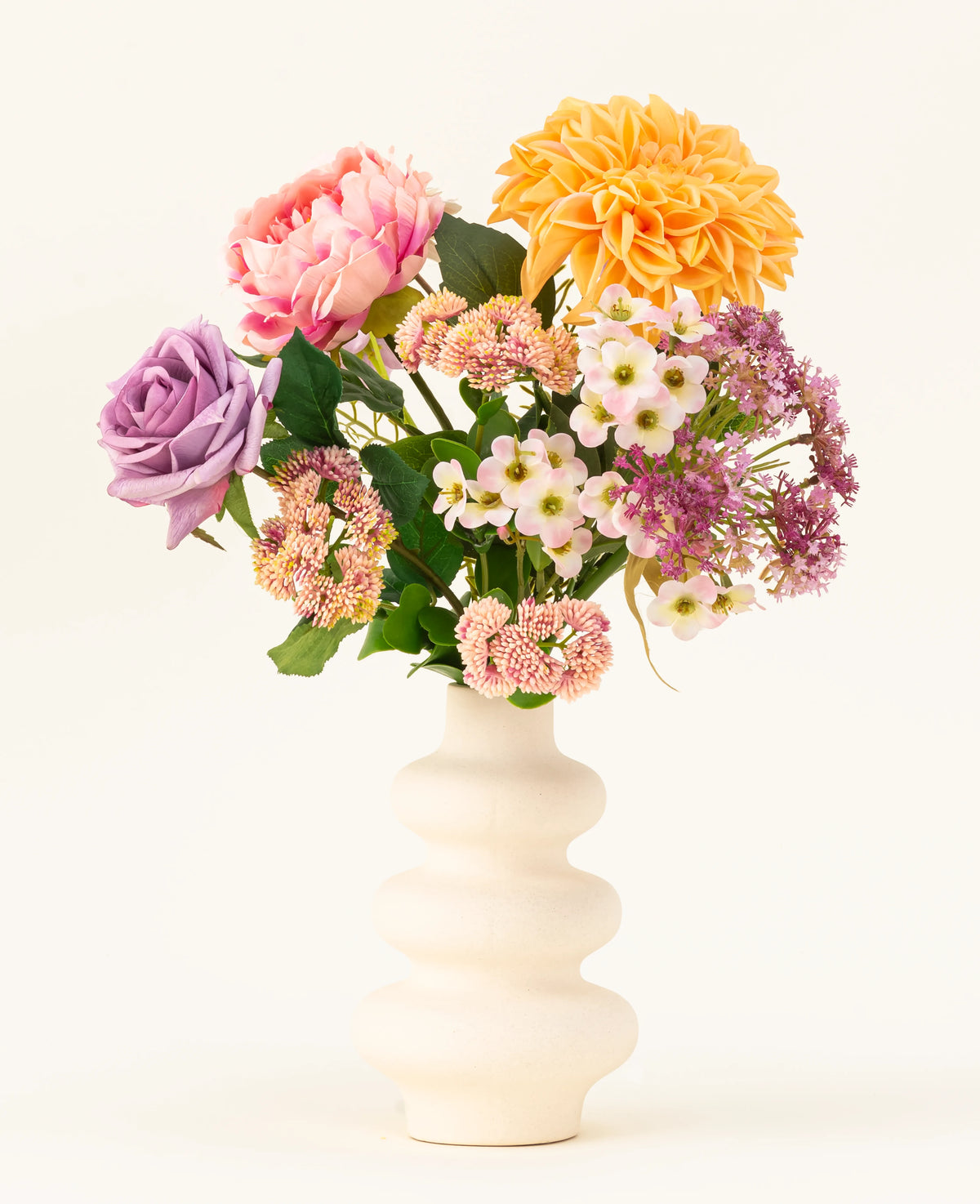 A white corrugated ceramic vase displays the Peach Delight silk artificial flower bouquet, featuring a yellow dahlia, pink peony, purple rose and several small white and pink flowers against a simple light background.