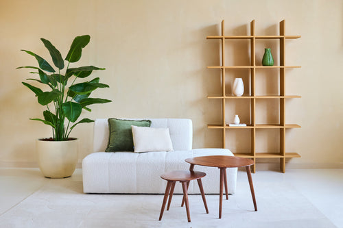 Minimalist living room with a white sofa, two cushions, wooden nesting tables and a large Artificial Plant Strelitzia Real Touch Deluxe 180 cm for a tropical atmosphere. A modern open wooden bookshelf with vases stands against the beige wall.
