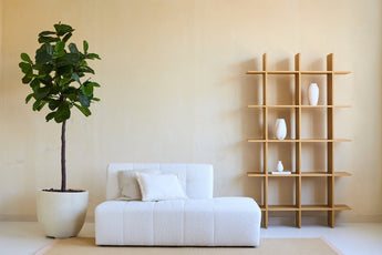 Minimalistische woonkamer met een witte bank, twee kussens en een Kunstplant Ficus Tabaksplant Deluxe 180 cm naast een open houten plank met witte vazen, alles tegen een beige muur en op een licht vloerkleed.