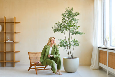 A woman in a green suit smiles as she sits on a wooden chair next to the Artificial Ruscus 180 cm plant in a bright, modern room with beige walls, large windows and minimalist decor.