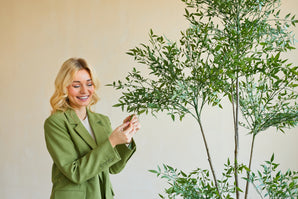 A smiling woman in a green blazer stands indoors and gently touches the leaves of an Artificial Ruscus 150 cm plant with thin branches and lush green foliage, standing out against a plain, light-colored background.