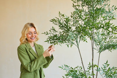 A smiling woman in a green blazer stands indoors and gently touches the leaves of an Artificial Ruscus 150 cm plant with thin branches and lush green foliage, standing out against a plain, light-colored background.
