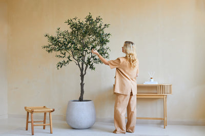 A woman in loose beige clothing stands next to a 200 cm Artificial Olive Tree and gently touches the leaves. Nearby, a wooden stool and a console table with books and a diffuser are against the beige wall.