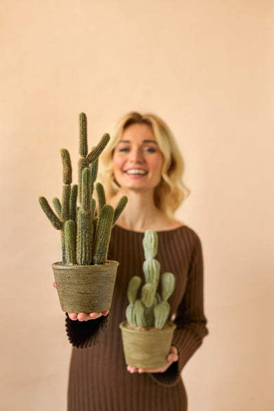 Een glimlachende vrouw in een bruine jurk houdt twee Kunst Cactus Cereus 35 cm planten in pot, een grote en een kleine, tegen een effen beige achtergrond.