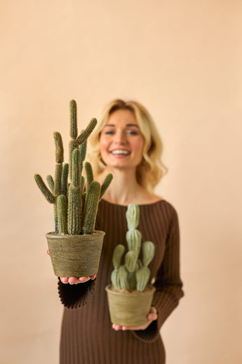A smiling woman in a brown dress holds two Artificial Cactus Cereus 35 cm potted plants, one large and one small, against a plain beige background.