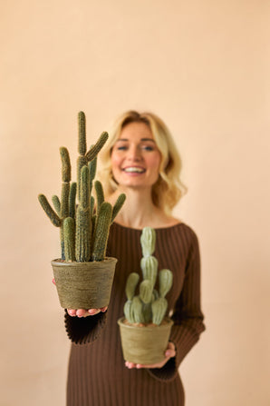 Een glimlachende vrouw in een bruine jurk houdt twee Kunst Cactus Cereus 35 cm planten in pot, een grote en een kleine, tegen een effen beige achtergrond.