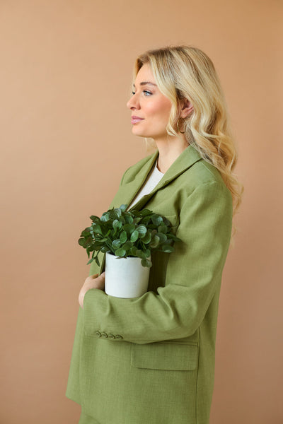 Een vrouw met lang blond haar in een lichtgroene blazer houdt de "Kunst vetplant 22 cm in sierpot" tegen haar borst en staat zijdelings voor een effen beige achtergrond.