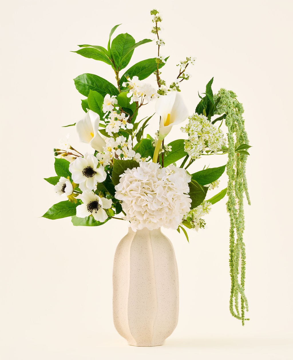 In a white ceramic vase is the 'Green Elegance' bouquet of silk artificial flowers, an arrangement of white hydrangeas, callas, anemones, green leaves and hanging greenery against a plain light background.