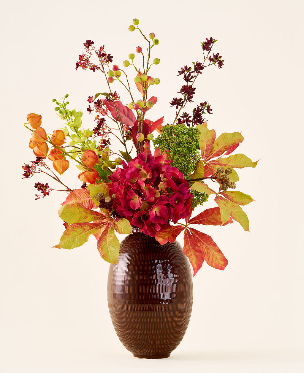 In a brown, ribbed vase is the bouquet “Silk Artificial Flowers Warm Amber”, with vibrant red, orange and green flowers and leaves on long stems, against a plain light background.