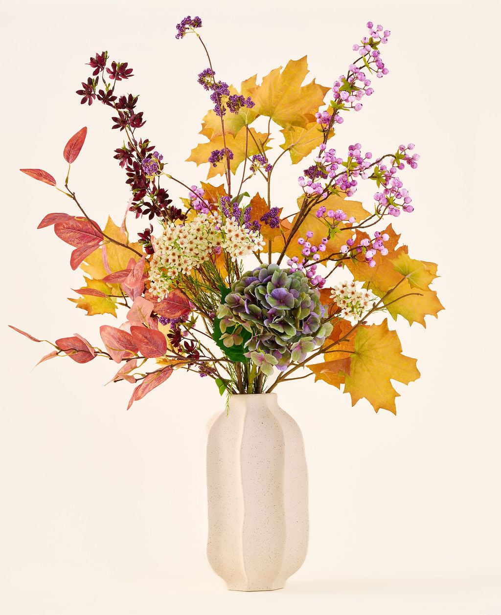 The 'Wild Amethyst' bouquet of silk artificial flowers, with autumn leaves, purple and white flowers and green hydrangeas, is in a white ceramic vase against a plain, light background.