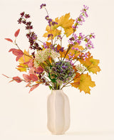 The 'Wild Amethyst' bouquet of silk artificial flowers, with autumn leaves, purple and white flowers and green hydrangeas, is in a white ceramic vase against a plain, light background.