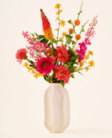 In a white ceramic vase is the 'Summer Blush' bouquet of silk artificial flowers, consisting of pink, yellow and orange flowers with green leaves, against a plain light background.