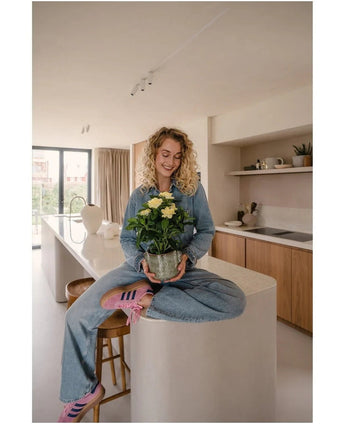 Een vrouw met krullend blond haar, gekleed in spijkerbroek en gestreepte sokken, zit glimlachend op het aanrecht terwijl ze de Kunstplant Gardenia UV 39 cm vasthoudt – een potplant met gele bloemen. Op de achtergrond is een moderne keuken met houten kasten en planken te zien.