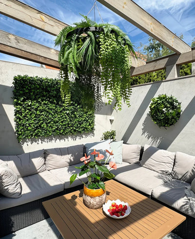 A sunny terrace with beige seating furniture with cushions and a wooden table, decorated with a pineapple vase with pink flowers and a bowl of strawberries. Green wall plants provide a lush interior, supplemented with the artificial hanging plant fern UV 60 cm in black ring as a striking hanging accent.