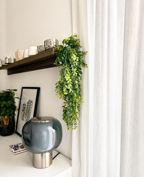 Een moderne woonruimte met een grijze glazen lamp, ingelijste kunst, een potplant en een houten plank met kopjes en de Kunst-hangplant ‘Pannenkoek’ (61 cm), waarvan de ranken naast witte gordijnen hangen.
