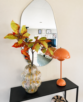 In a wavy mirror above a black console table there is a glass vase with Kunsttak Chestnut 91 cm red/green and an orange mushroom lamp. The mirror reflects a living room with framed wall art.