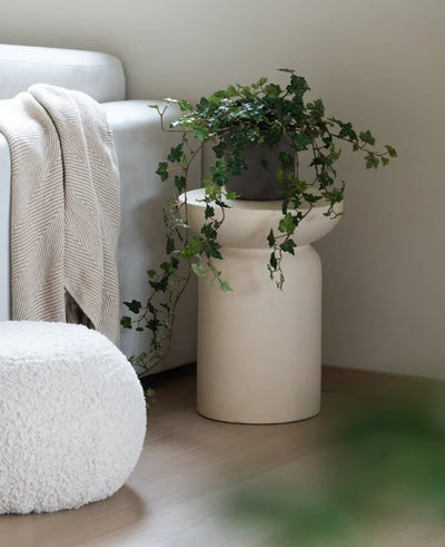 An Artificial Ivy Deluxe Green 90 cm stands in a beige vase on a wooden floor next to a light gray sofa with a cream-colored plaid and a white structured pouffe.