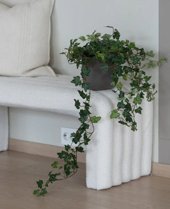 The Artificial Ivy Deluxe Green 90 cm, with its lush hanging tendrils, stands on a white structured bench next to a cream-colored cushion in a minimalist room with light wooden floors.