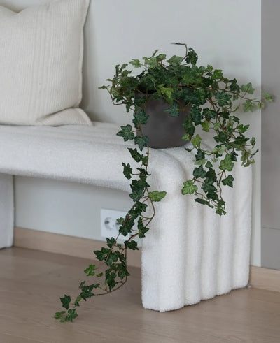 The Artificial Ivy Deluxe Green 90 cm, with its lush hanging tendrils, stands on a white structured bench next to a cream-colored cushion in a minimalist room with light wooden floors.