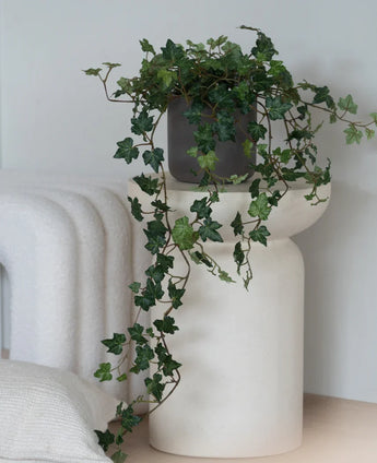 An Artificial Ivy Deluxe Green, a 90 cm hanging ivy in a gray pot, stands on a modern, white, sculptural base next to a folded white blanket and a white structured cushion.