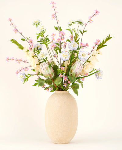 In een vaas met beige structuur staat het Zijden kunstbloemen boeket Lots of Love XL, een handgebonden boeket van roze, witte en blauwe zijden bloemen met groene bladeren en lange stelen tegen een lichte achtergrond.