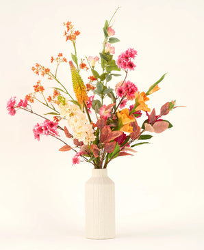 In een vaas met witte structuur staat het Zijden kunstbloemen boeket Wild Peach met kleurrijke kunstbloemen - roze, oranje, geel en wit - en groene en roodachtige bladeren tegen een effen lichte achtergrond.
