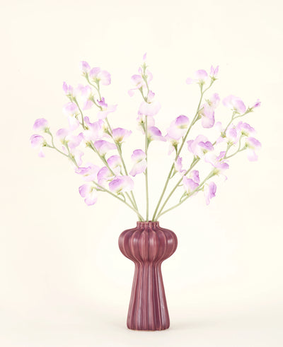 Het Zijden kunstbloemen boeket Erwt lila heeft lange stelen van delicate, lichtpaarse bloemen en groene bladeren in een vaas van mauve keramiek met verticale richels, tegen een effen, gebroken witte achtergrond.