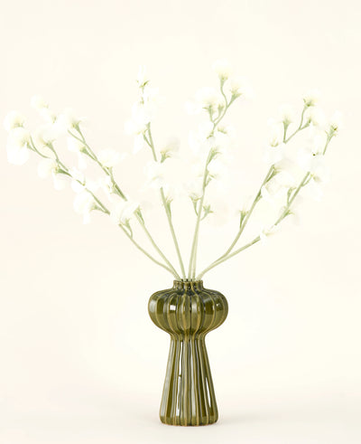 Een groene geribbelde glazen vaas bevat het Zijden kunstbloemen boeket Erwt crème met delicate witte zijden bloemen en slanke stelen, tegen een effen gebroken witte achtergrond.