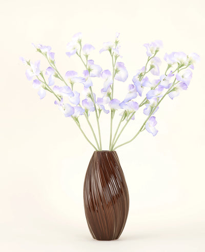 Een bruine, geribbelde vaas bevat het Zijden kunstbloemen boeket Erwt lavendel, met delicate bloemblaadjes en groene stelen tegen een effen gebroken witte achtergrond voor een subtiele cottage sfeer.