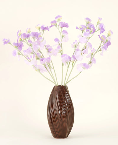 Een bruine vaas met textuur bevat het Zijden kunstbloemen boeket Erwt paars-lichtpaarse zijden bloemen met groenachtige stelen tegen een effen, lichte achtergrond.