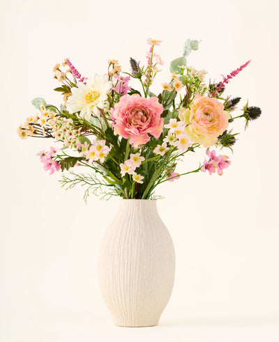 In een witte keramische vaas staat het boeket ‘Backyard Blossom’ van Zijden kunstbloemen, bestaande uit een prachtig arrangement van roze rozen, witte madeliefjes en kleine gele bloemetjes, vervaardigd uit zijden bloemen, aangevuld met weelderig groen blad tegen een effen lichte achtergrond.
