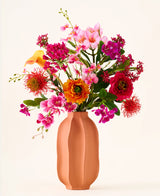 In a peach-colored vase is the bouquet "Tropical Sunset" of silk artificial flowers, consisting of a lively mix of pink, orange, yellow and red flowers against a plain, light background.
