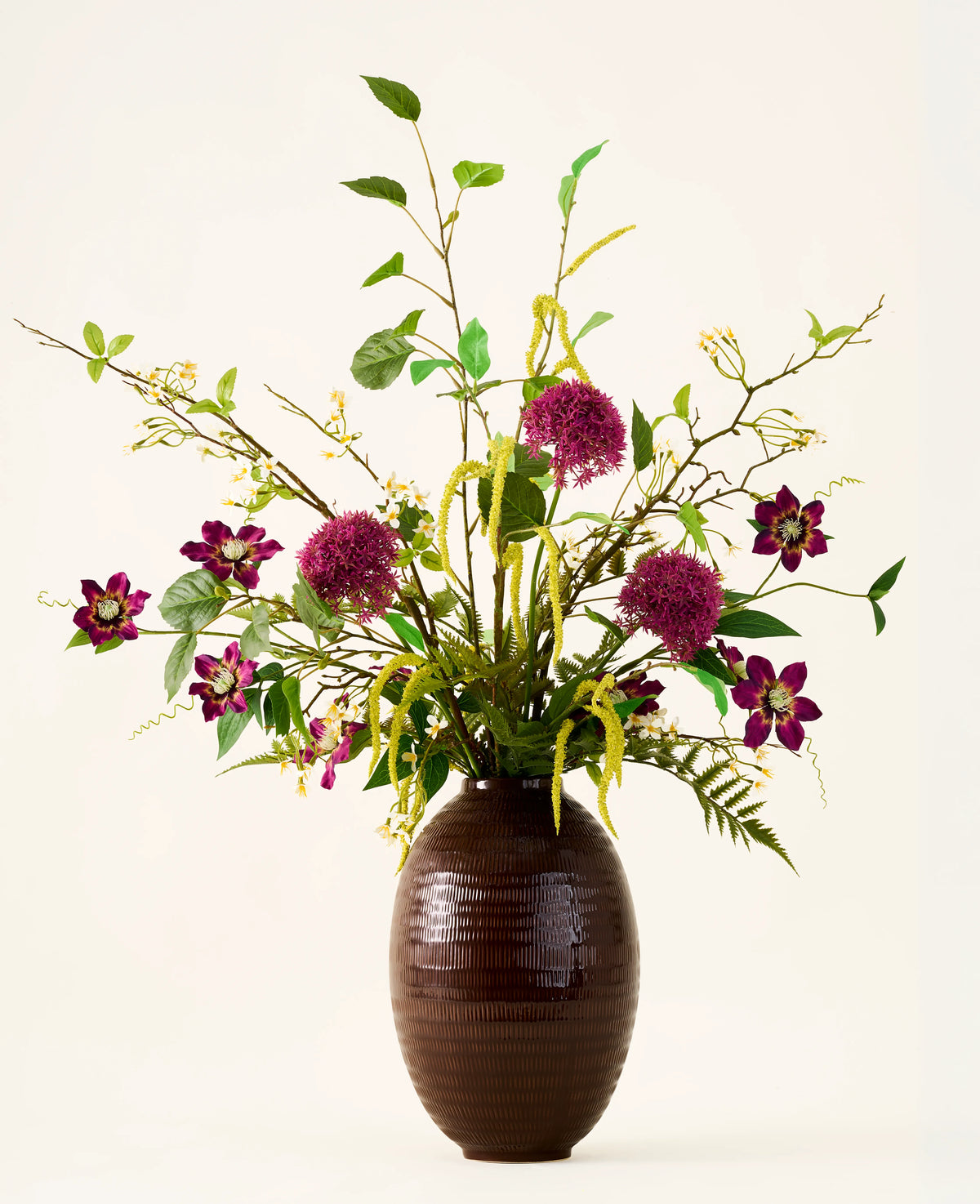 In a brown, ribbed vase is the bouquet "Woodland Charm" of silk artificial flowers, with purple alliums, dark pink flowers, yellow twigs and green leaves against a plain, light background.