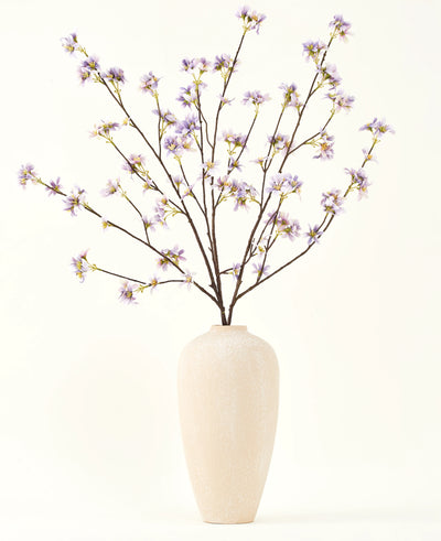 Een hoge, crèmekleurige vaas van keramiek bevat een aantal dunne takken met de tekst Zijden kunstbloemen boeket Kunstbloementak paars, tegen een effen, lichte achtergrond.