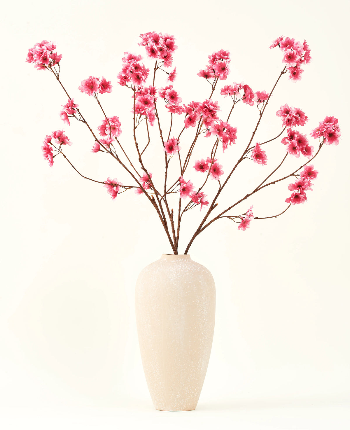 The Silk artificial flower bouquet Cherry blossom fuchsia 114 cm adds a vibrant pop of color to your interior with its striking branches and fuchsia blossoms, beautifully displayed in a beige ceramic vase against a plain off-white background.