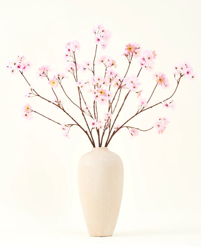 Het Zijden kunstbloemen boeket Kersenbloesem roze 114 cm heeft lange, dunne takken met trossen lichtroze bloemen in een beige vaas van keramiek, die een opvallend contrast vormen tegen de effen witte achtergrond.