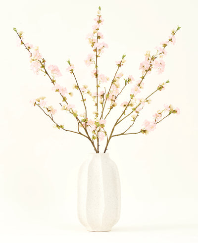Het Zijden kunstbloemen boeket Kersenbloesem lichtroze 94 cm heeft delicate roze kunstmatige kersenbloesems in een witte keramieken vaas, die elegantie toevoegt tegen een effen lichte achtergrond.