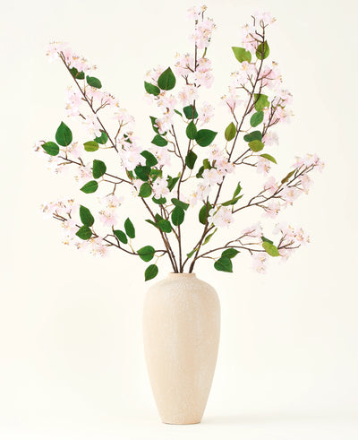 Een beige vaas van keramiek bevat verschillende lange takken met het Zijden kunstbloemen boeket Perzikbloesem roze en groene bladeren, tegen een effen witte achtergrond - perfect voor een kunstbloemen interieur.