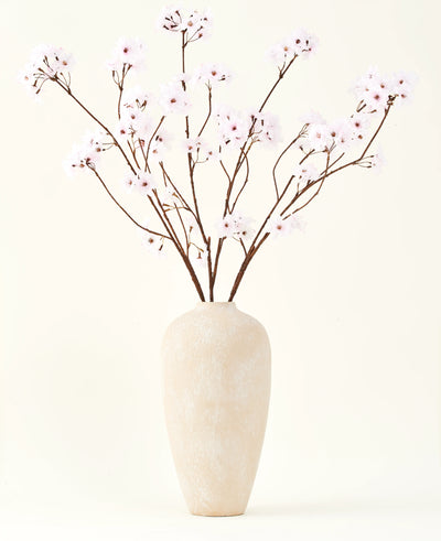 De Zijden kunstbloemen boeket Kersenbloesem lichtroze 114 cm staat in een hoge vaas van beige keramiek met lange, dunne takken en delicate lichtroze bloemen, waardoor een sereen kunstbloemen accent ontstaat tegen een effen achtergrond.