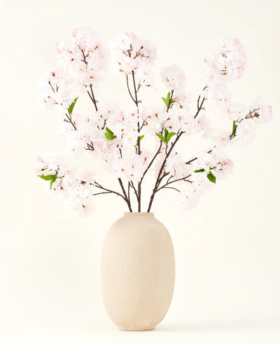 Het Zijden kunstbloemen boeket Kersenbloesem blush 119 cm toont delicate, lichtroze kersenbloesem met groene bladeren in een beige vaas, ideaal als kunstbloemen decoratie tegen een lichte achtergrond.