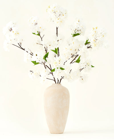 Het Zijden kunstbloemen boeket Kersenbloesem crème 119 cm heeft elegante bruine takken met zacht witte nepbloesem en groene bladeren, weergegeven in een eenvoudige beige vaas tegen een effen lichte achtergrond.