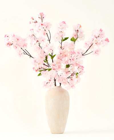 De Zijden kunstbloemen boeket Kersenbloesem roze 119 cm, gerangschikt in een beige keramische vaas met delicate groene bladeren, creëert een sereen interieuraccent tegen een eenvoudige lichte achtergrond.