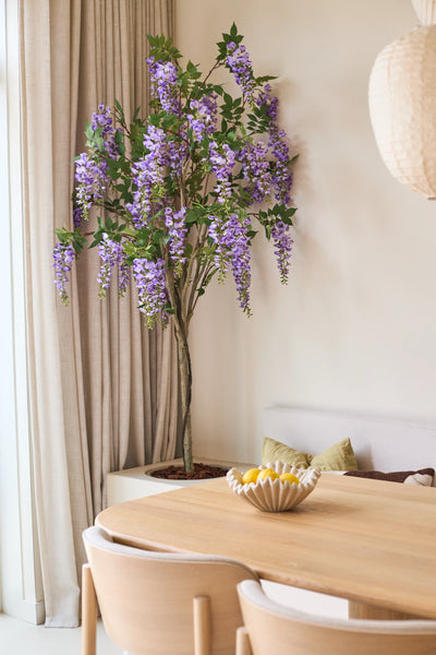 An artificial Wisteria plant measuring 180 cm with lilac-blue flower clusters stands near a window with beige curtains, next to a wooden table with a bowl of lemons and a bench with green cushions.