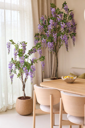 Two Artificial Wisteria plants of 150 cm, with their overhanging purple flowers, stand next to a wooden dining table with light chairs. Soft natural light shines through the translucent curtains behind the plants.