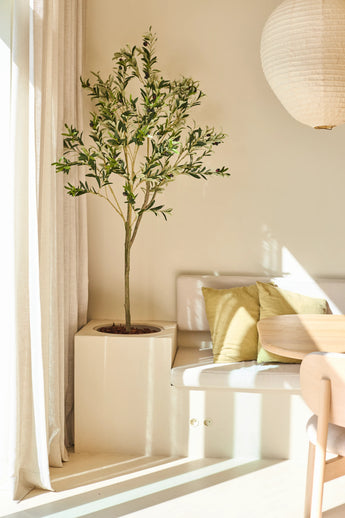 The 150 cm Artificial Olive Tree stands next to a white bench with green and beige cushions, near a round wooden table and a hanging paper lamp in a sunlit, minimalist room.